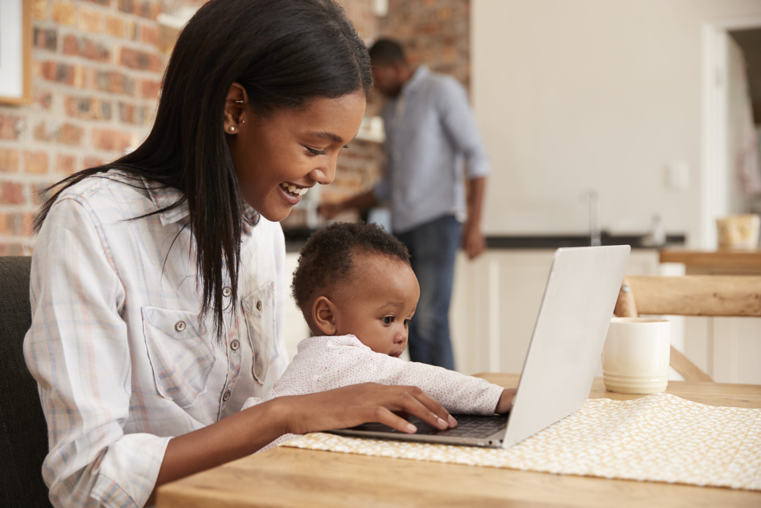 Mother And Baby Daughter Use Laptop As Father Prepares Meal